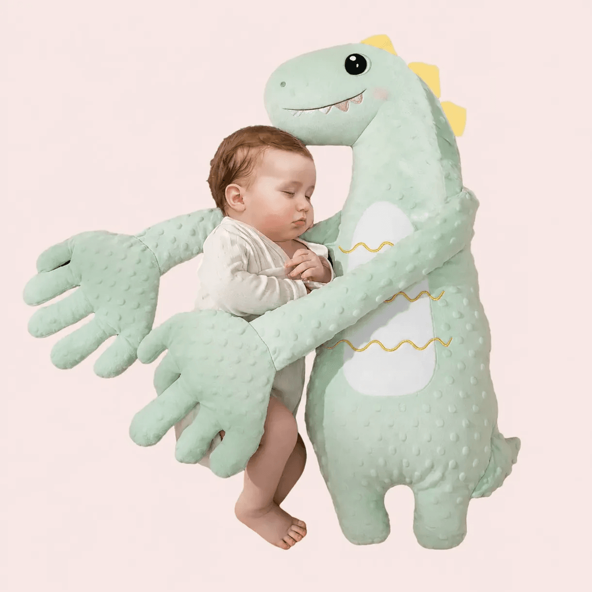 A light green textured plush dinosaur toy with large padded hands laying beside a sleeping baby on a light background.