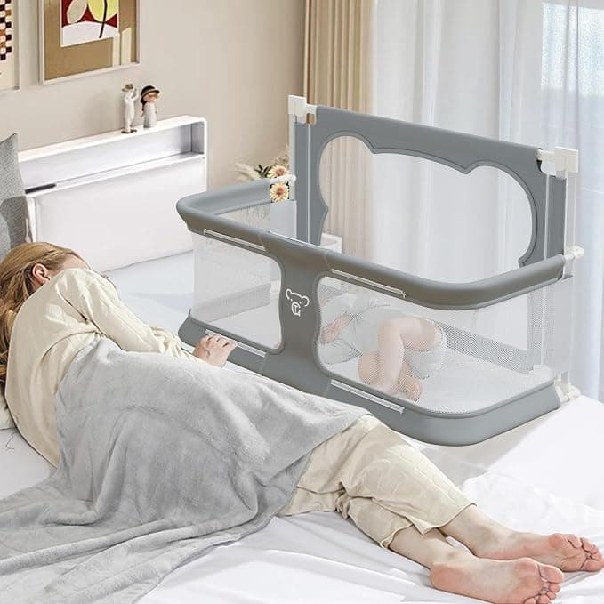 A gray mesh bedside sleeper attached to an adult bed, with a sleeping baby inside next to a resting adult.