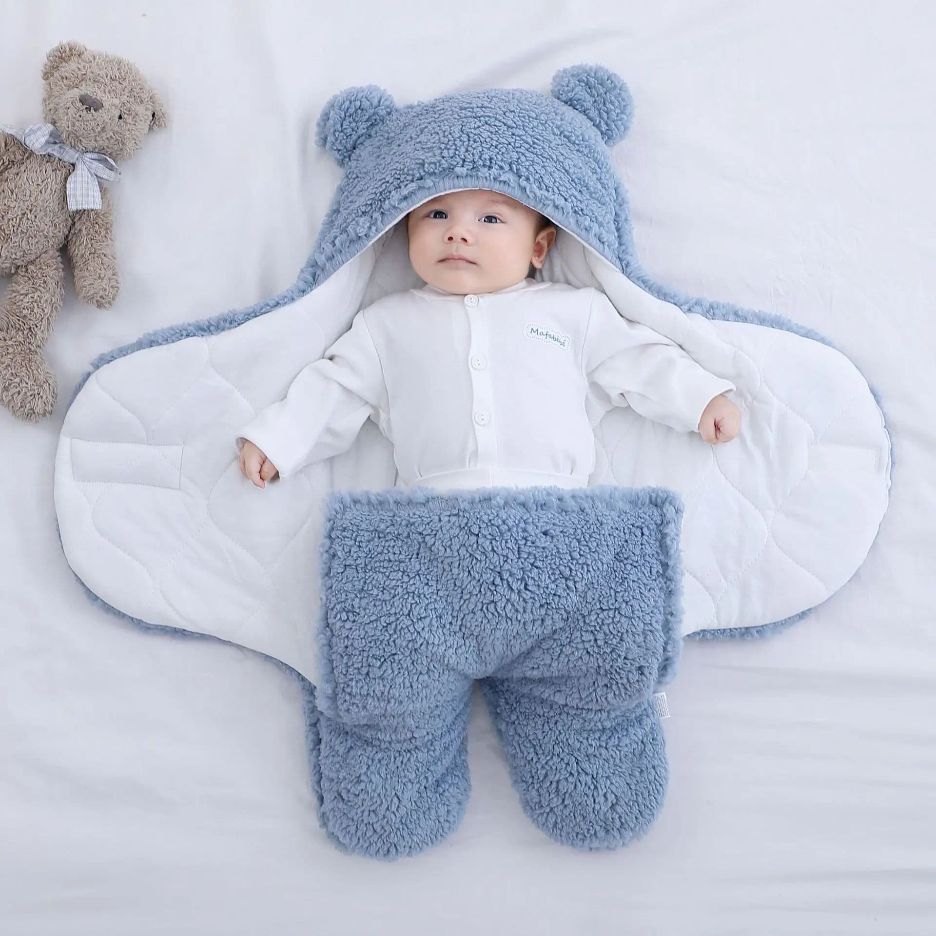 A baby resting inside a blue plush swaddle wrap with bear ears on the hood and separate leg pockets, lined with white quilted fabric.