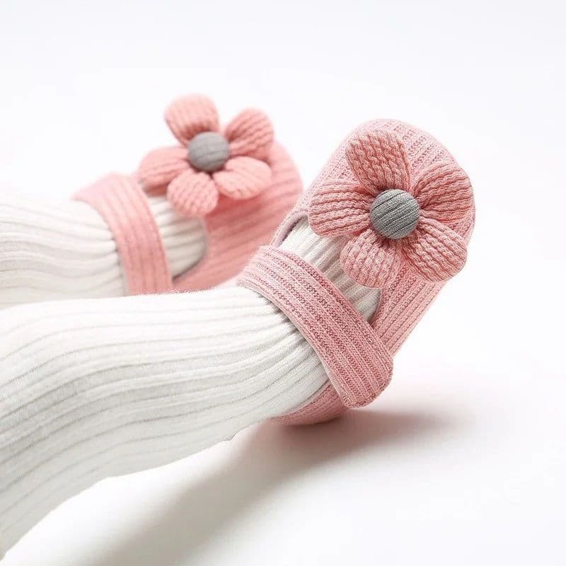 Pink knitted baby prewalker shoes with decorative flower details on top, shown worn over white ribbed tights.