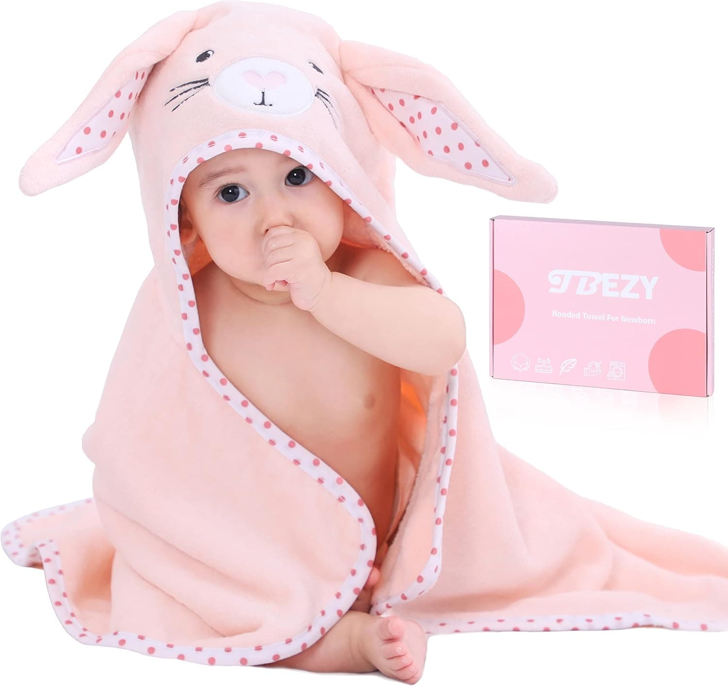 A baby wrapped in a light pink hooded towel with bunny ears and a polka dot trim next to its product packaging box.