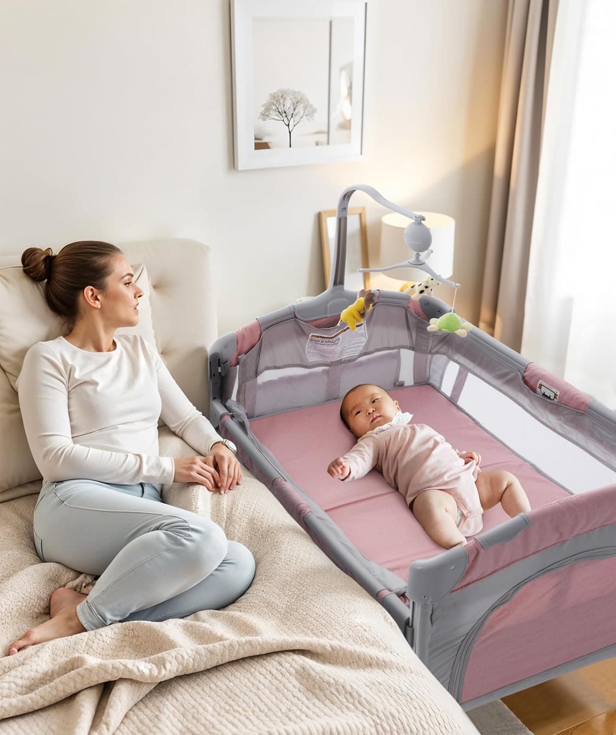 An infant rests inside a pink and grey bedside crib with mesh sides next to an adult bed, underneath a small hanging mobile attachment.